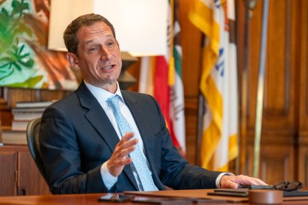 A man in a suit and tie sits at a desk with flags in the background, gesturing with one hand while speaking.