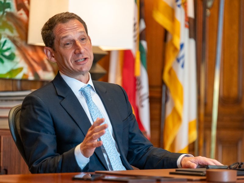 A man in a suit and tie sits at a desk with flags in the background, gesturing with one hand while speaking.
