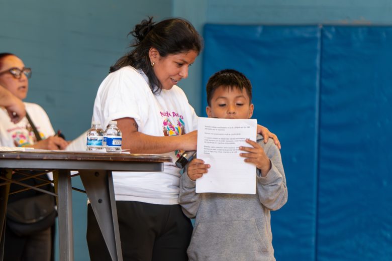 An immigrant woman helps a boy read from a paper while standing indoors near a table with water bottles; another person is in the background.
