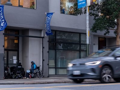 A blur of a gray car drives past a city street. A person in a wheelchair and another seated are near a building entrance with blue banners. Trees and buildings line the sidewalk.