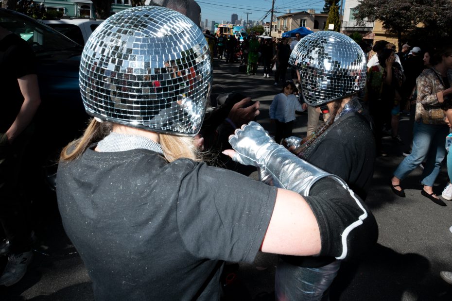 Two people wearing mirrored disco ball helmets and shiny silver gloves interact at an outdoor street event with several other people in the background.