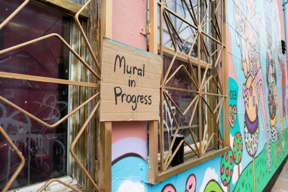 A cardboard sign reading "Mural in Progress" is taped to a window with metal bars beside a partially painted, colorful mural on an exterior wall.