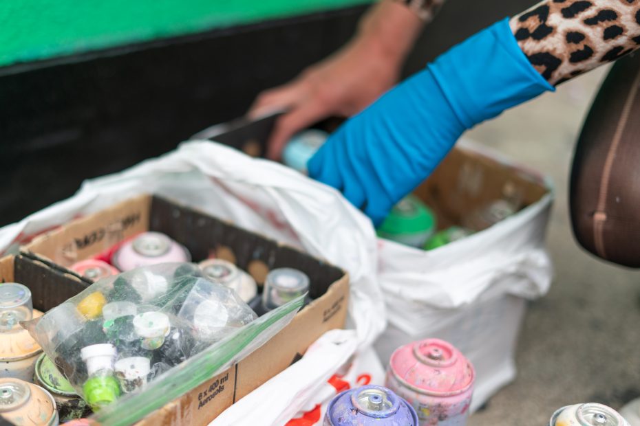 A person wearing a blue glove handles spray paint cans stored in cardboard boxes lined with white plastic bags.