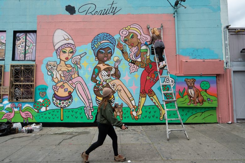 A person on a ladder paints a colorful mural of three stylized women with animals on a building, while another person walks by on the sidewalk.