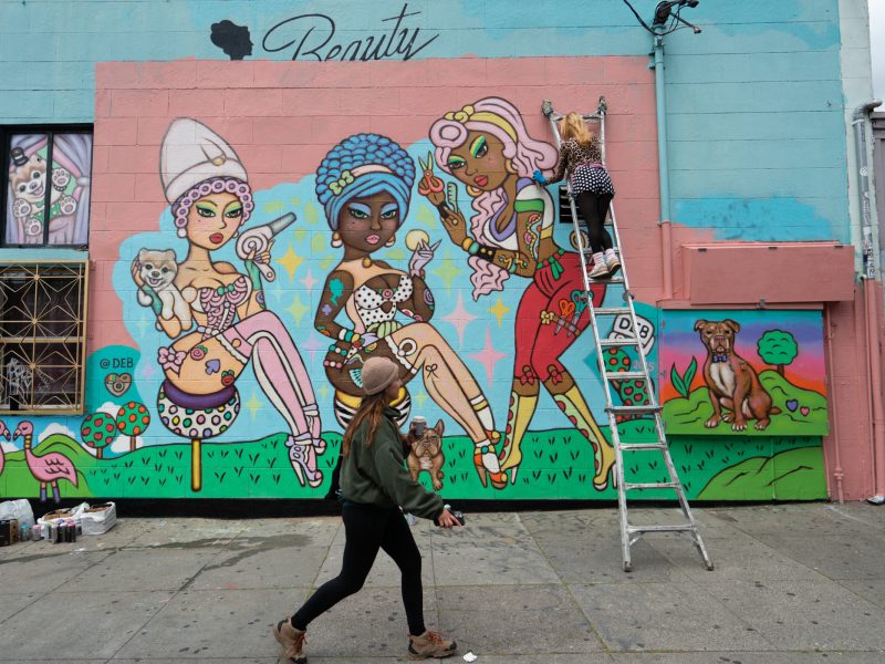 A person on a ladder paints a colorful mural of three stylized women with animals on a building, while another person walks by on the sidewalk.