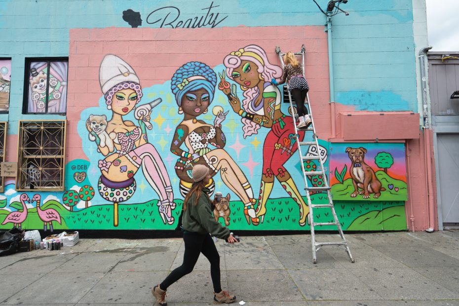 A person on a ladder paints a colorful mural of stylized women and animals on a building while another person walks by on the sidewalk.