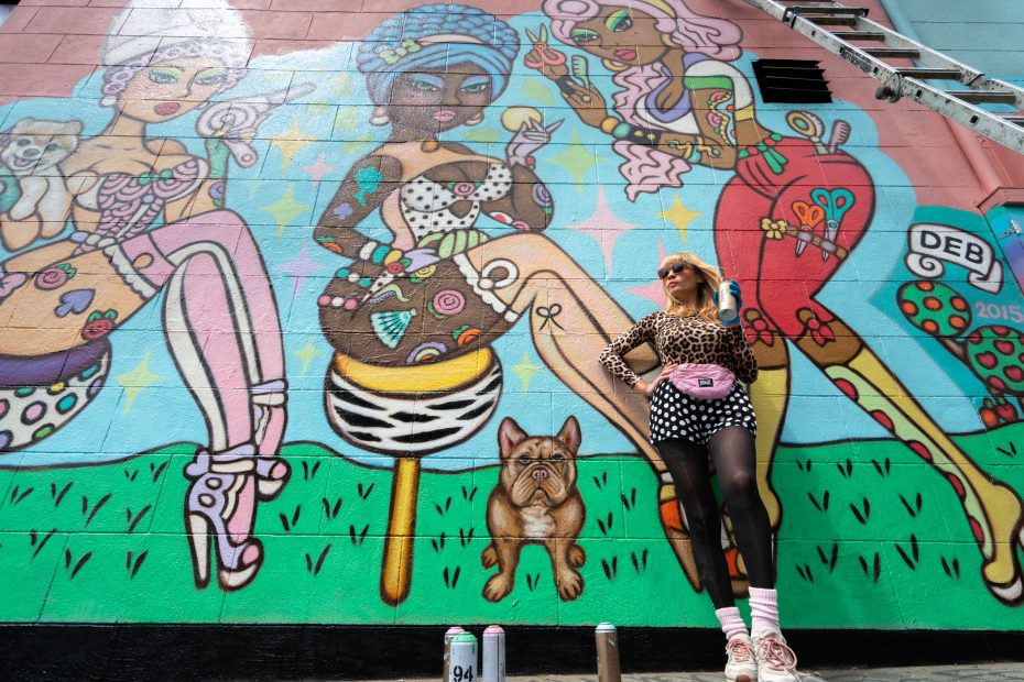 A person in patterned clothing and sunglasses poses in front of a colorful mural featuring three stylized women, a dog, and various patterns on an outdoor wall.
