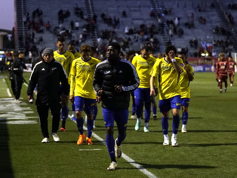 A group of soccer players in yellow jerseys and blue shorts walk off the field together as spectators sit in the stadium stands.