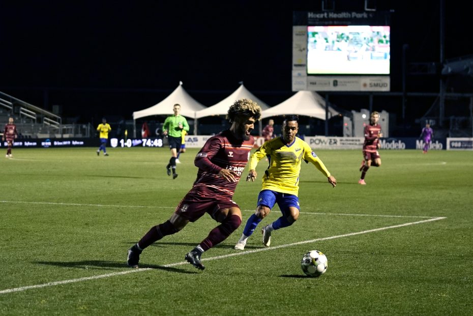 A soccer player in a maroon uniform dribbles the ball while being pursued by a player in a yellow jersey during a night match.