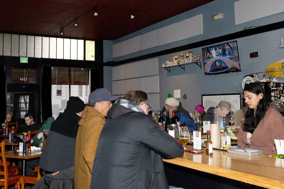 People sit at a bar and tables inside a restaurant, some eating and drinking, with a TV on the wall showing a sports game.