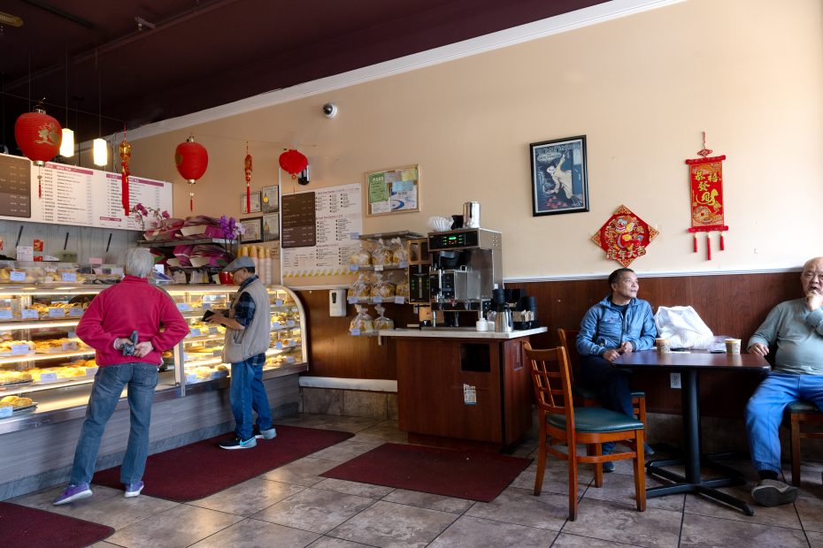 A Chinese bakery with customers at the counter and seated at tables; display cases with pastries, red lanterns, and festive decorations are visible.