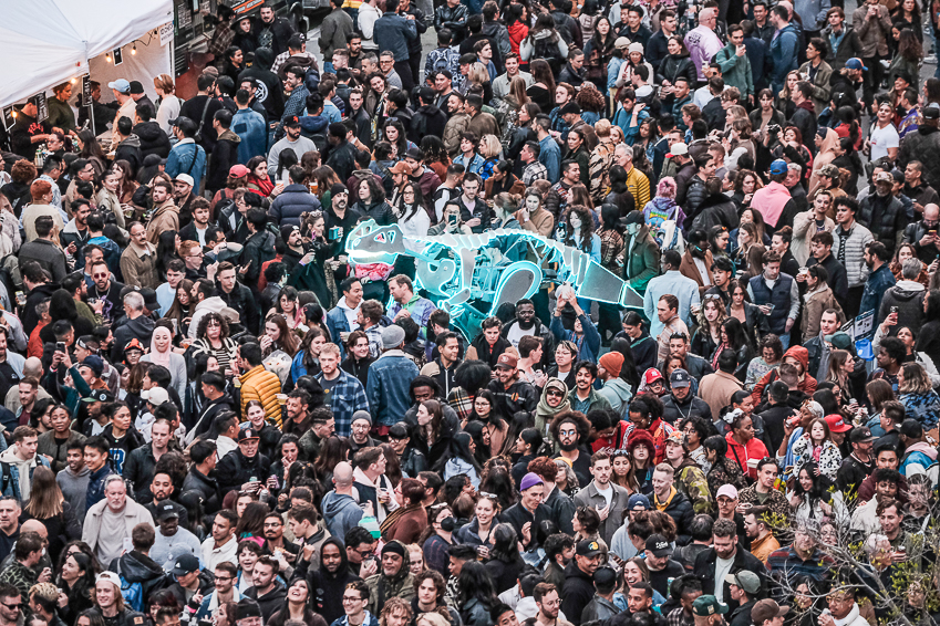 A large crowd gathers downtown, with a glowing, inflatable dragon among them. Many people are looking up, possibly at a mesmerizing performance or display.