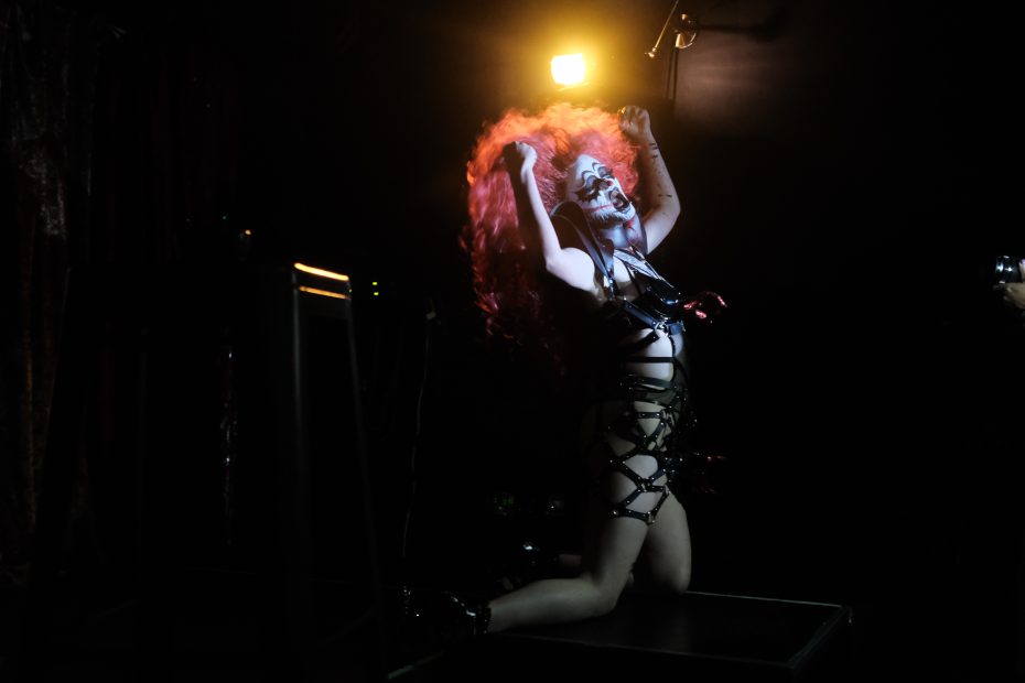 Performer in dramatic makeup and outfit kneels on stage under spotlight, hands raised, with vibrant, long red hair flowing.