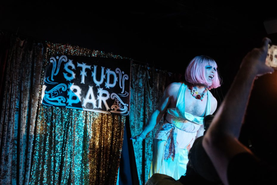 Performer with pink wig and colorful attire on stage at Stud Bar, engaging with the audience.