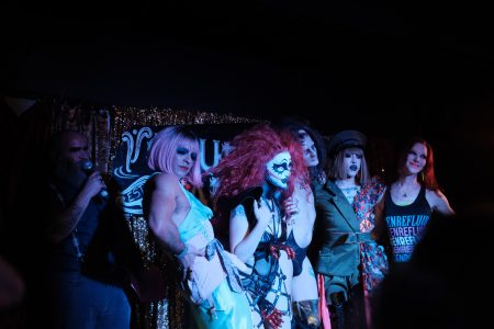 Five performers in colorful, theatrical costumes and makeup stand on stage under dim lighting. A person with a microphone is speaking to them from the side.