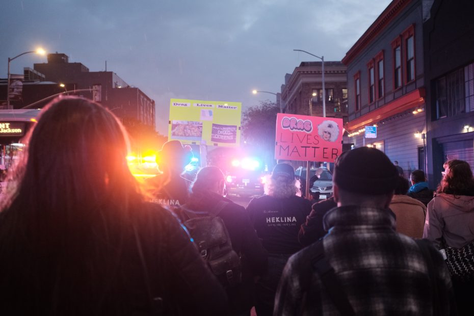 A group of people with signs, including one stating "Drag Lives Matter," gather near police vehicles with flashing lights on a city street at dusk.