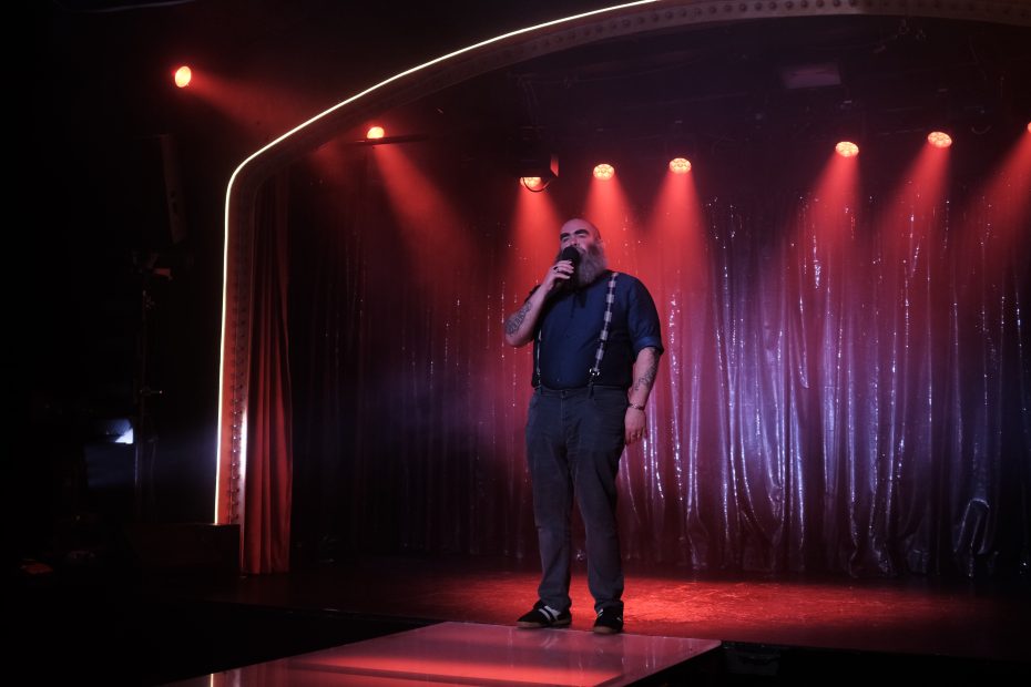 A bearded man wearing suspenders stands on a stage with a microphone, under red lighting and in front of shimmering curtains.