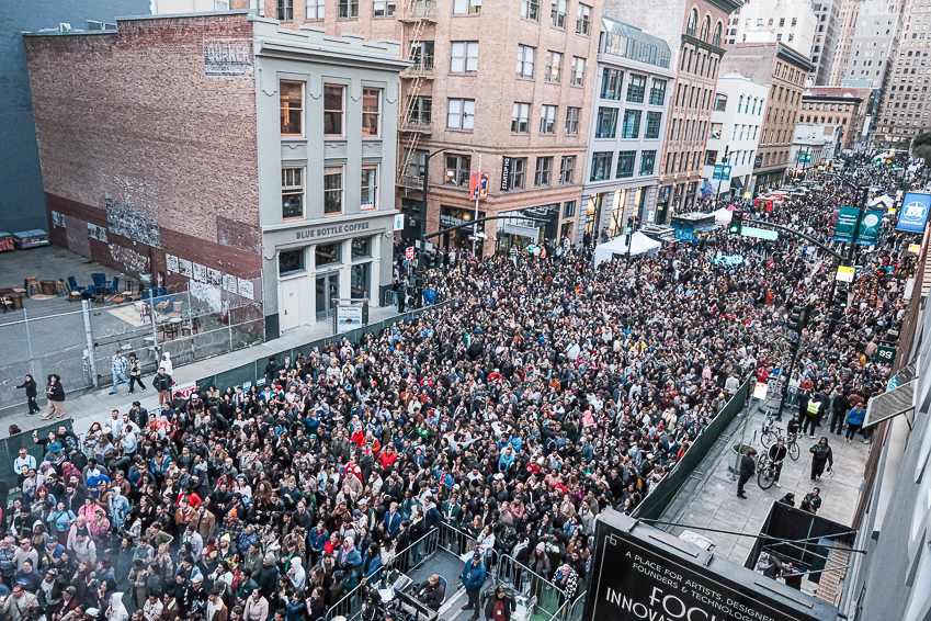 A large crowd gathers on a bustling downtown city street, bordered by towering buildings. Many people stand closely together, filling the entire street and sidewalks with energy and excitement.