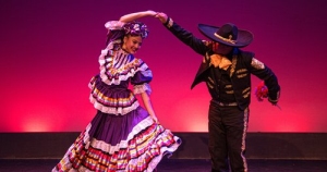 A woman in a colorful traditional dress and a man in a charro outfit perform a folkloric dance on stage with a purple and pink background.