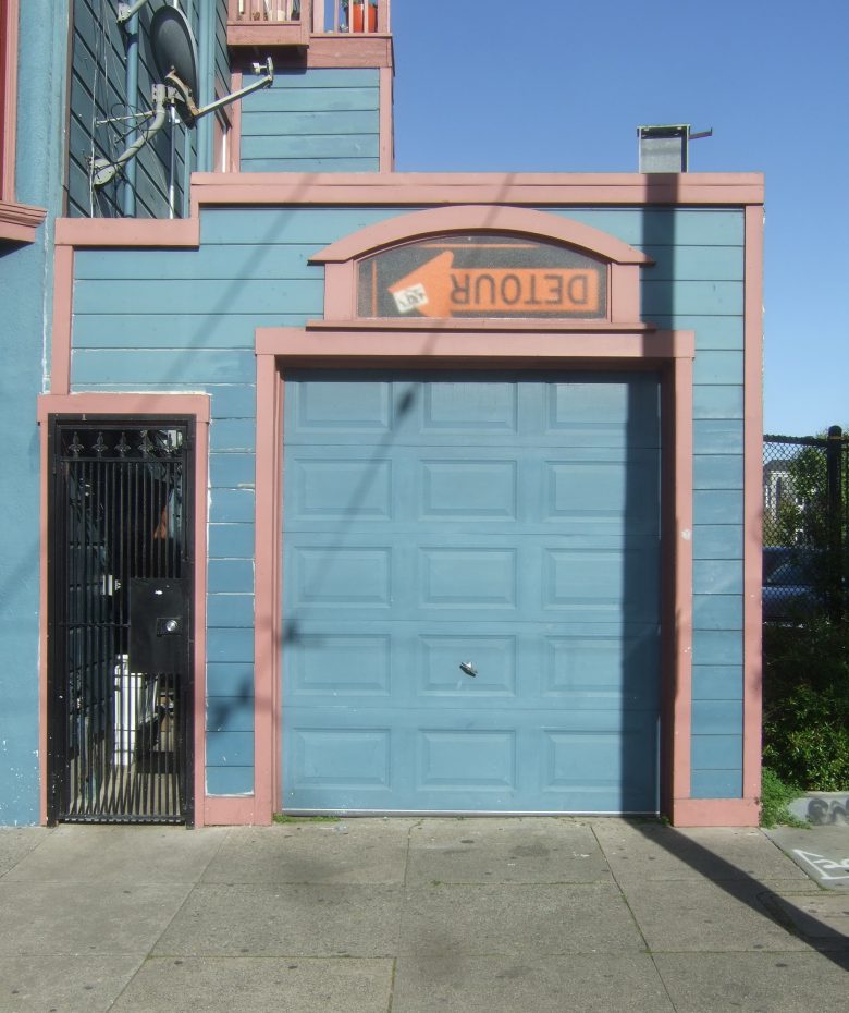 Blue garage door in a colorful building with a humorous "Detour" sign placed upside down above it. Shadow of a pole is cast on the door.