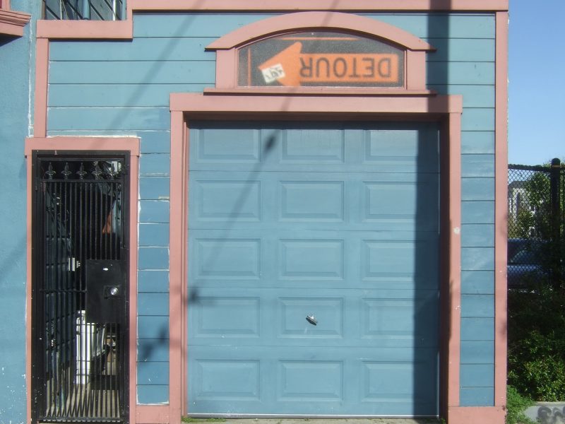 Blue garage door in a colorful building with a humorous "Detour" sign placed upside down above it. Shadow of a pole is cast on the door.