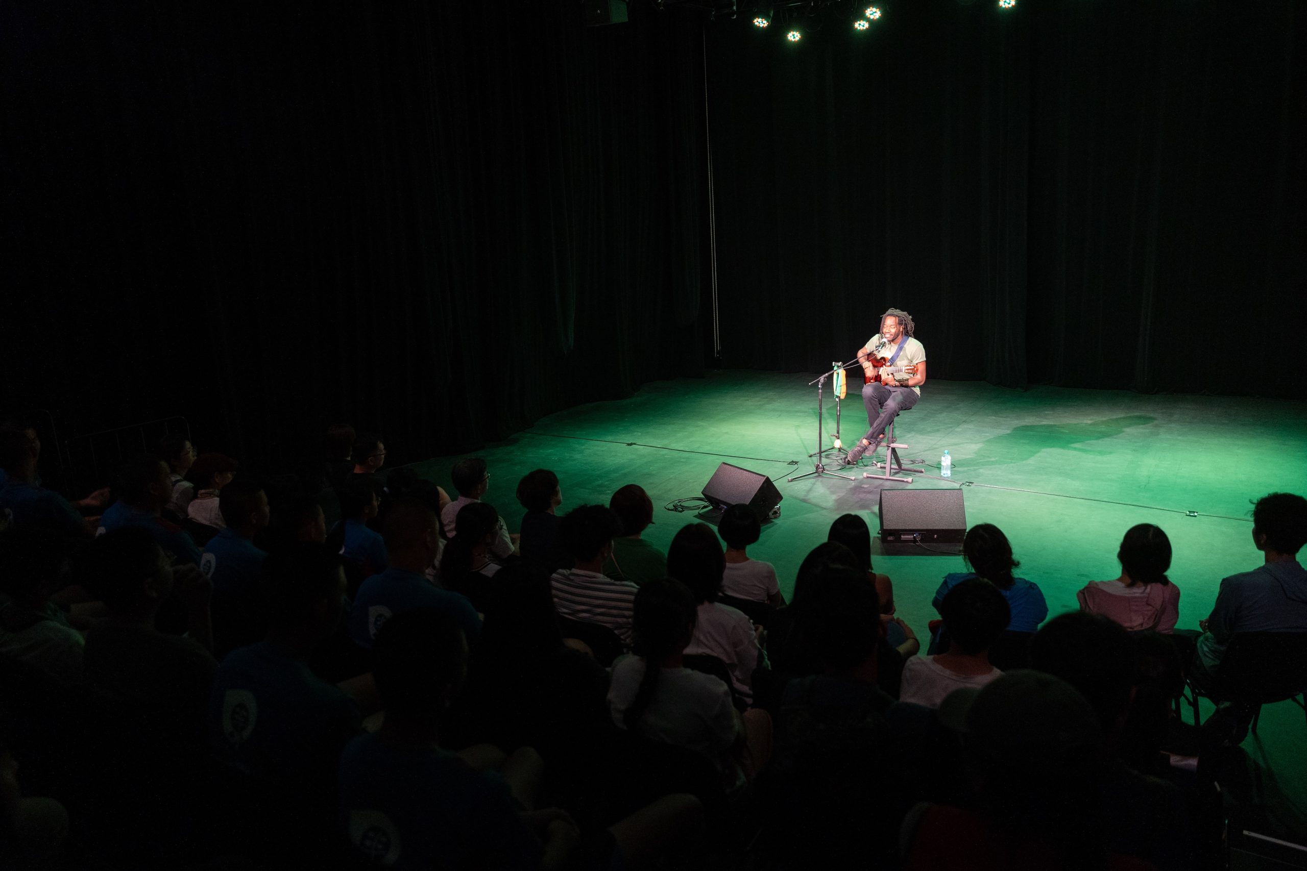 A solo musician sits on stage playing guitar and singing under stage lights, while an audience watches in a dark theater.