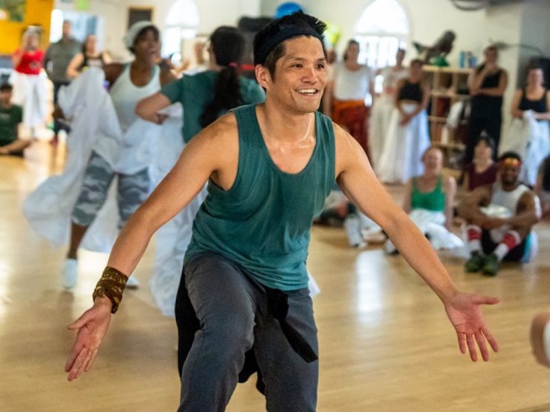 A man in a green tank top and dark pants dances joyfully in a studio, surrounded by other people participating in a group dance class.