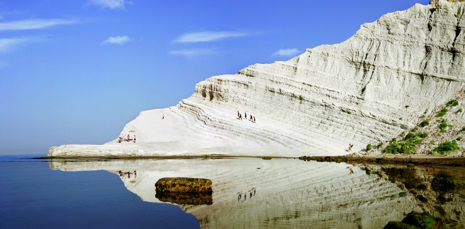 A large white limestone cliff slopes down to calm water, reflecting the rock formation and a few people climbing the natural steps.