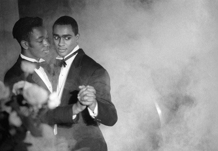 Two men in tuxedos slow dance together, holding hands, surrounded by mist or fog.