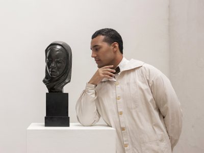 A person in a light-colored coat stands next to a sculpture of a woman's head on a pedestal, gazing thoughtfully at the artwork in a minimalist gallery space.