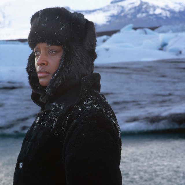 A person in a black fur hat and coat stands near icy water with snow-covered mountains and glaciers in the background.
