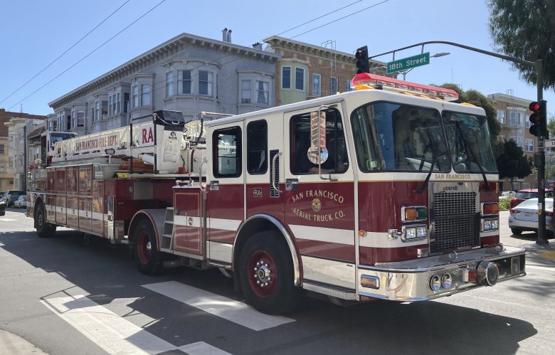 A San Francisco fire truck is parked at an intersection in sunlight. Residential buildings are visible in the background.