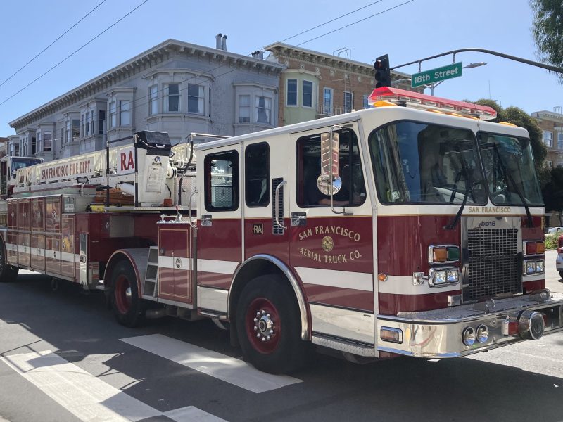 A San Francisco fire truck is parked at an intersection in sunlight. Residential buildings are visible in the background.