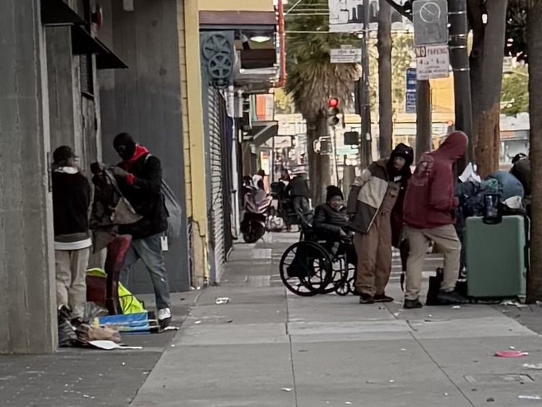 Several people, including one in a wheelchair, are gathered on a city sidewalk near buildings, with belongings and litter scattered around.