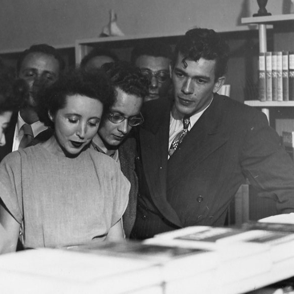 A group of people in formal attire closely examine books on a table in a library or bookstore setting.