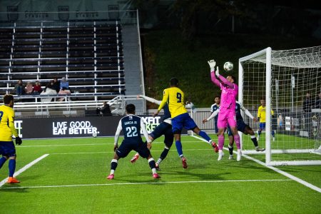 A soccer goalkeeper in pink jumps to block a ball near the goalpost as players from both teams attempt to score or defend.