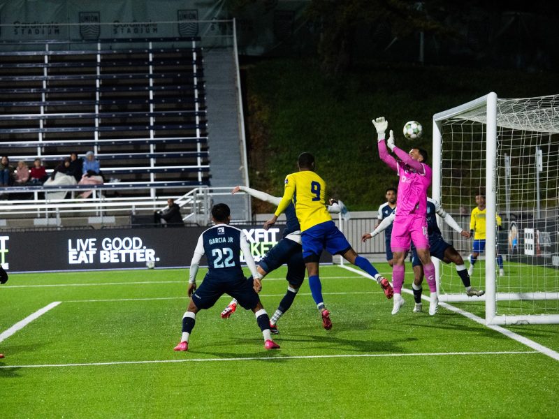 A soccer goalkeeper in pink jumps to block a ball near the goalpost as players from both teams attempt to score or defend.