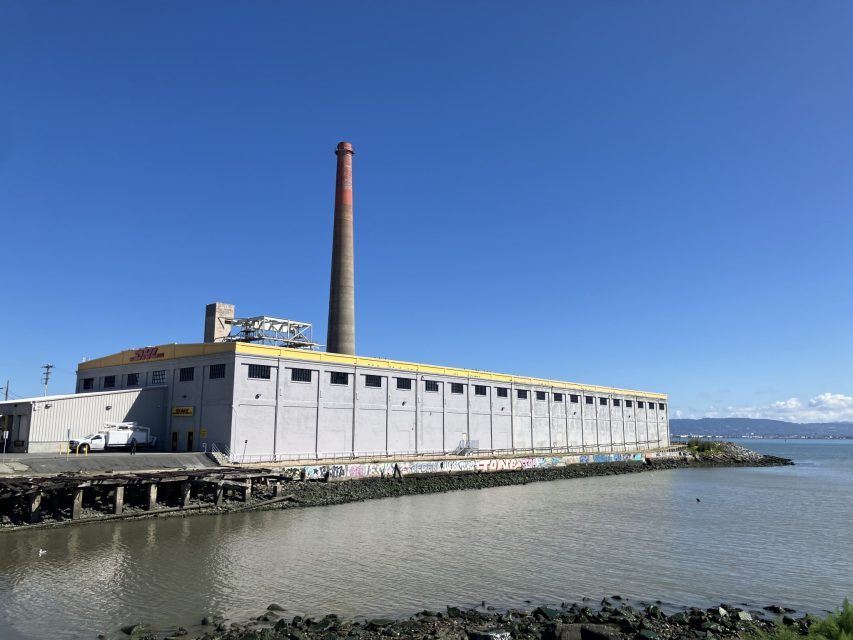 A large industrial building with a tall smokestack stands by the water, under a clear blue sky. Graffiti is visible on the lower part of the structure.