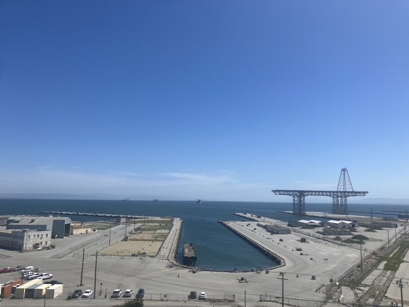 A waterfront industrial area with docks, empty lots, parked vehicles, and a large steel crane structure beside the water under a clear blue sky.