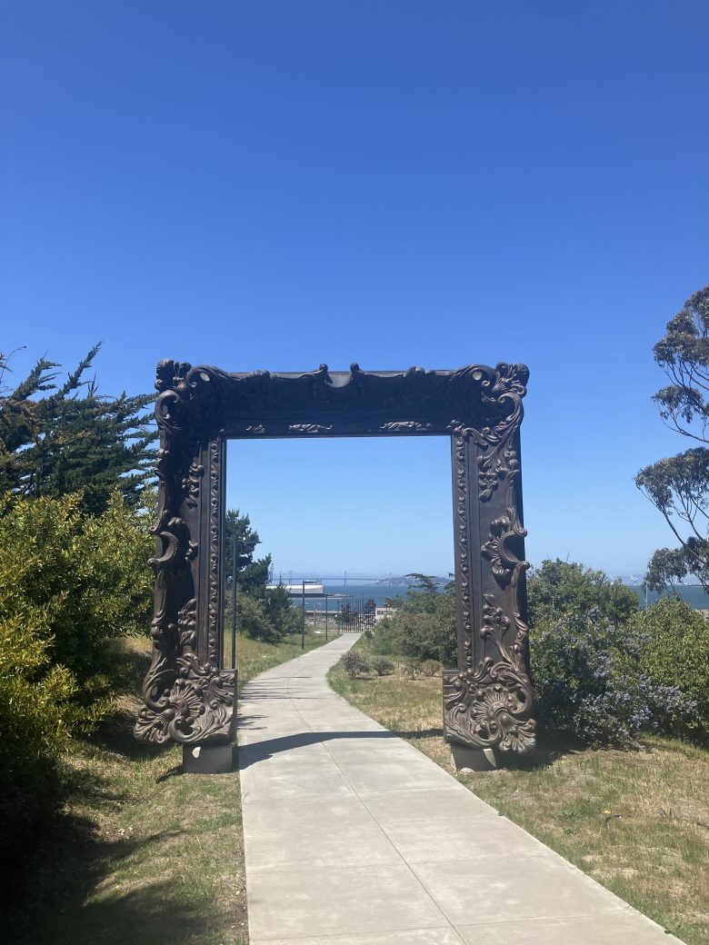 A decorative, oversized picture frame sculpture stands over a paved path, with trees and distant water visible under a clear blue sky.