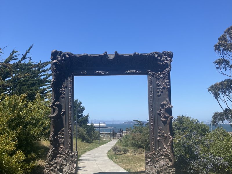 A decorative, oversized picture frame sculpture stands over a paved path, with trees and distant water visible under a clear blue sky.