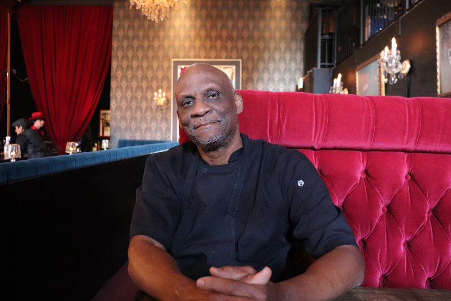 A man in a black chef coat sits on a red tufted bench in a dimly lit restaurant with patterned wallpaper and chandeliers.