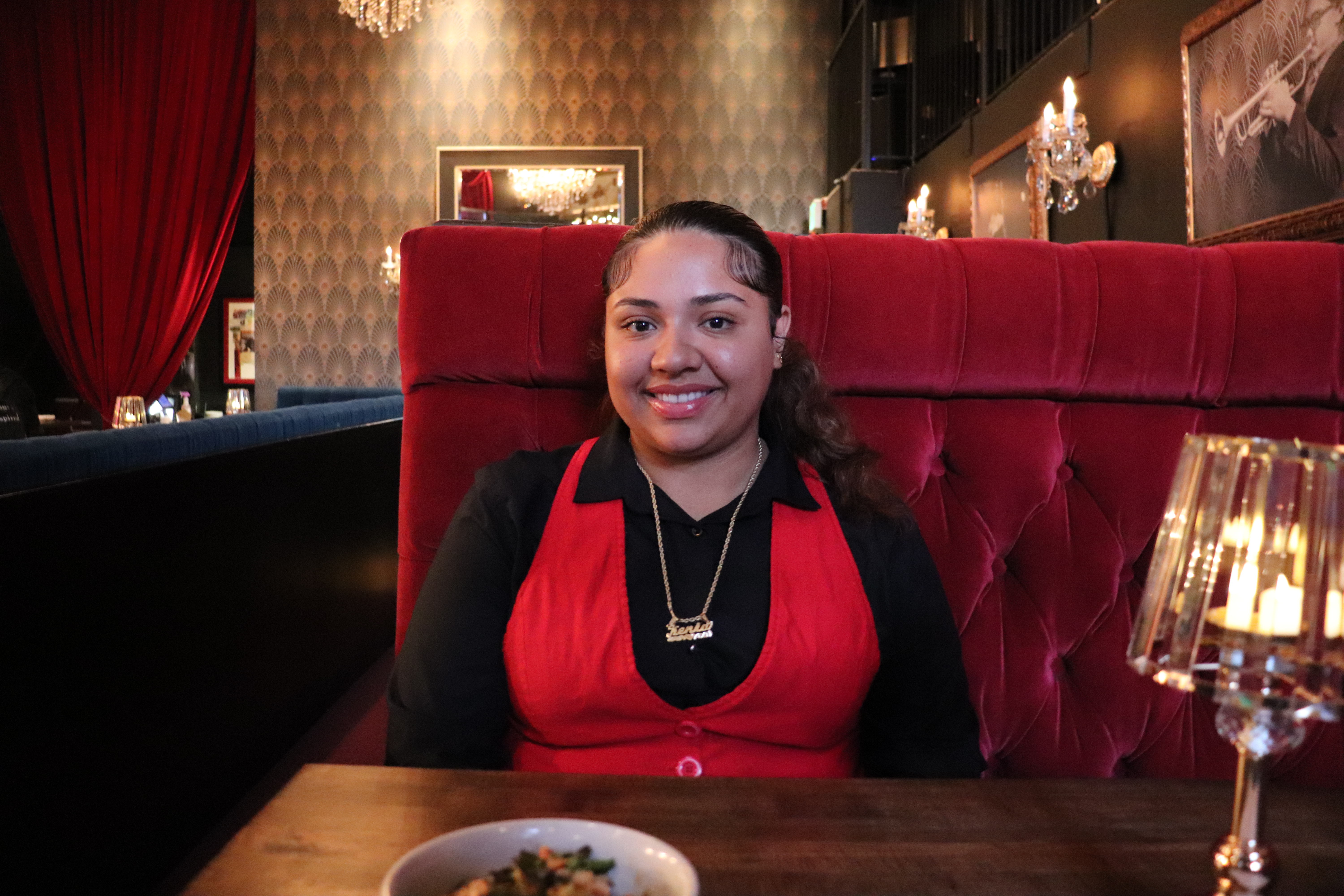 A woman in a red and black outfit sits at a wooden table in a dimly lit, elegant restaurant with chandeliers and red velvet decor. A bowl of food is on the table in front of her.