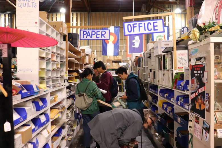 People browse shelves of office and paper supplies in a store aisle labeled "Paper" and "Office," surrounded by various stationery and books.