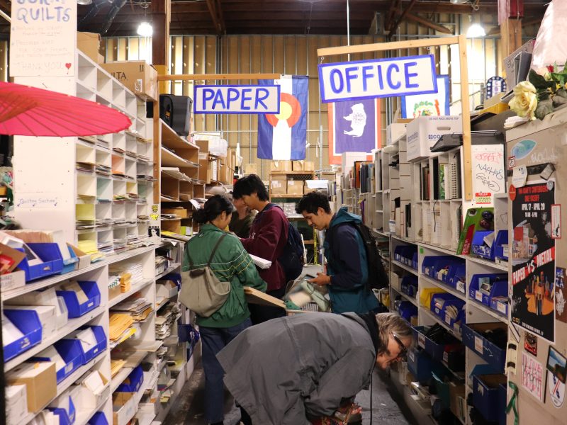 People browse shelves of office and paper supplies in a store aisle labeled "Paper" and "Office," surrounded by various stationery and books.