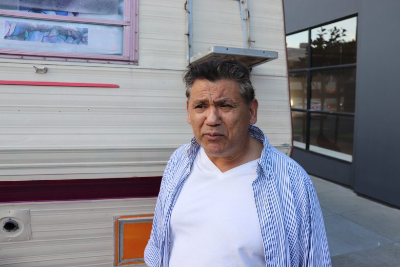 A man in a white shirt and striped overshirt stands in front of a camper trailer parked on a city street.