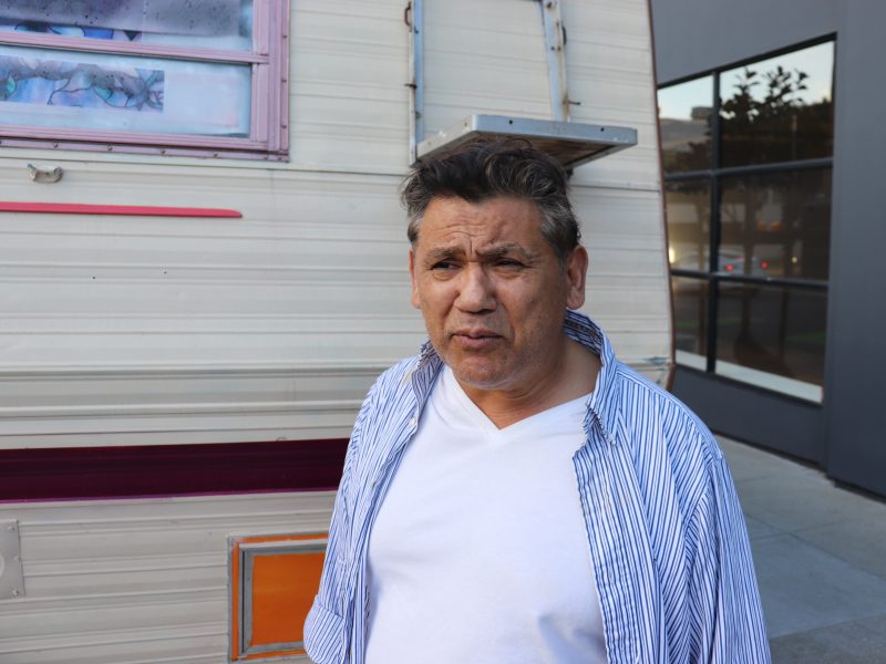 A man in a white shirt and striped overshirt stands in front of a camper trailer parked on a city street.