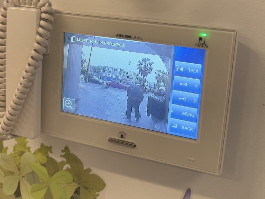 A security monitor screen shows a live video feed of a person standing near a gate outside, with buildings, palm trees, and a sidewalk visible in the background.
