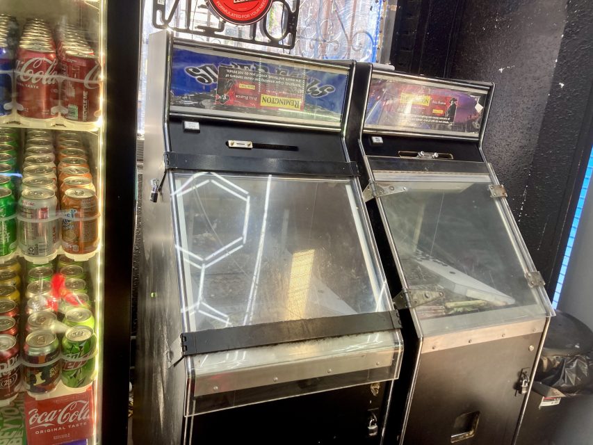 Two vintage pinball machines next to a refrigerated display filled with various soda cans, including Coca-Cola. The pinball machines have clear glass tops and black exteriors.