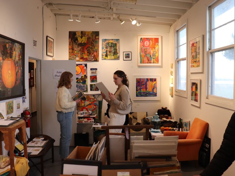 Two women stand in an art gallery, looking at artwork. Colorful paintings cover the walls, and bins with prints or art are in the foreground.
